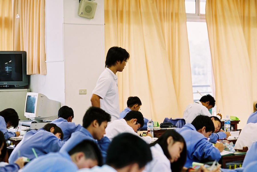 Man overlooks his students who have their heads down as they're taking a paper test.